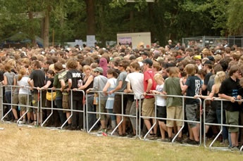 A row of fixed base cross tube police barriers are installed on the ground and a lot of people stand beside of it.